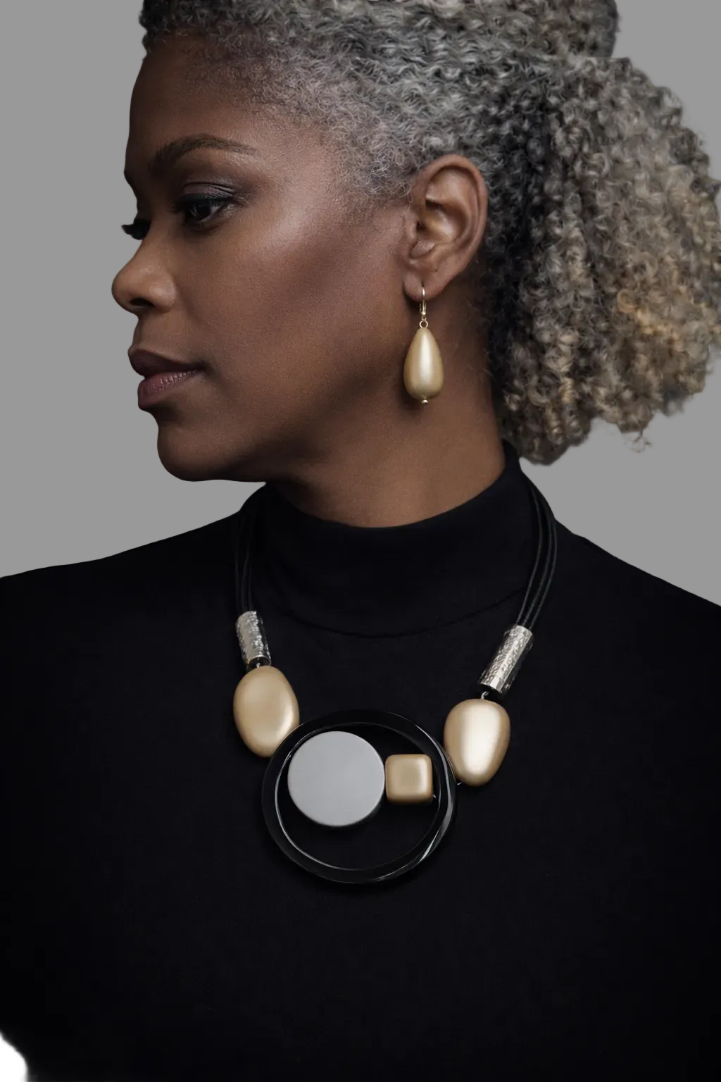 Woman wearing a black turtleneck and a unique necklace and earring set with gold and silver elements on a white background
