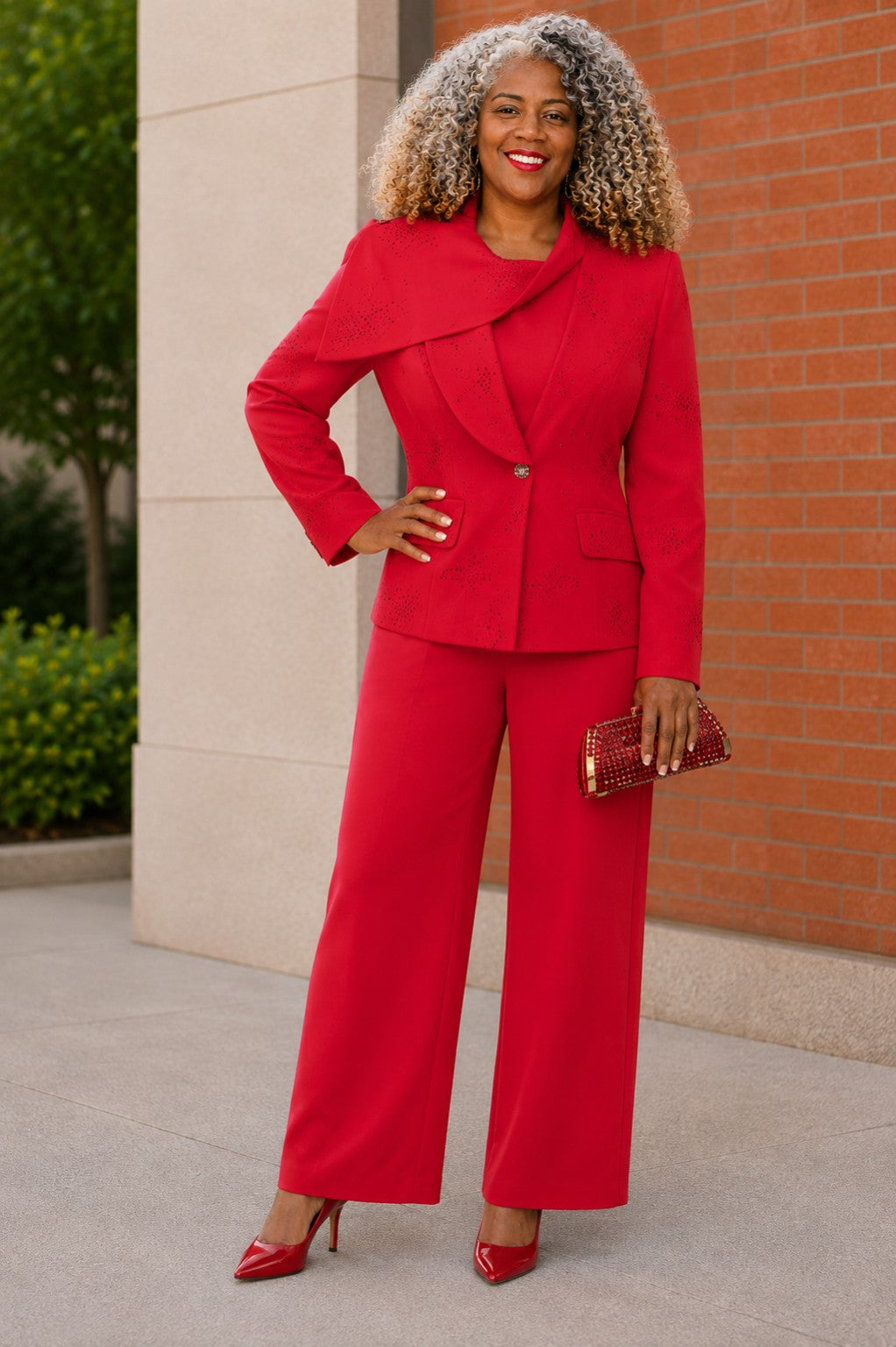 A woman wearing a red tailored pantsuit with a structured embellished jacket and wide-leg trousers, standing outdoors against a brick wall. She is holding a small metallic clutch and wearing red pointed heels.
