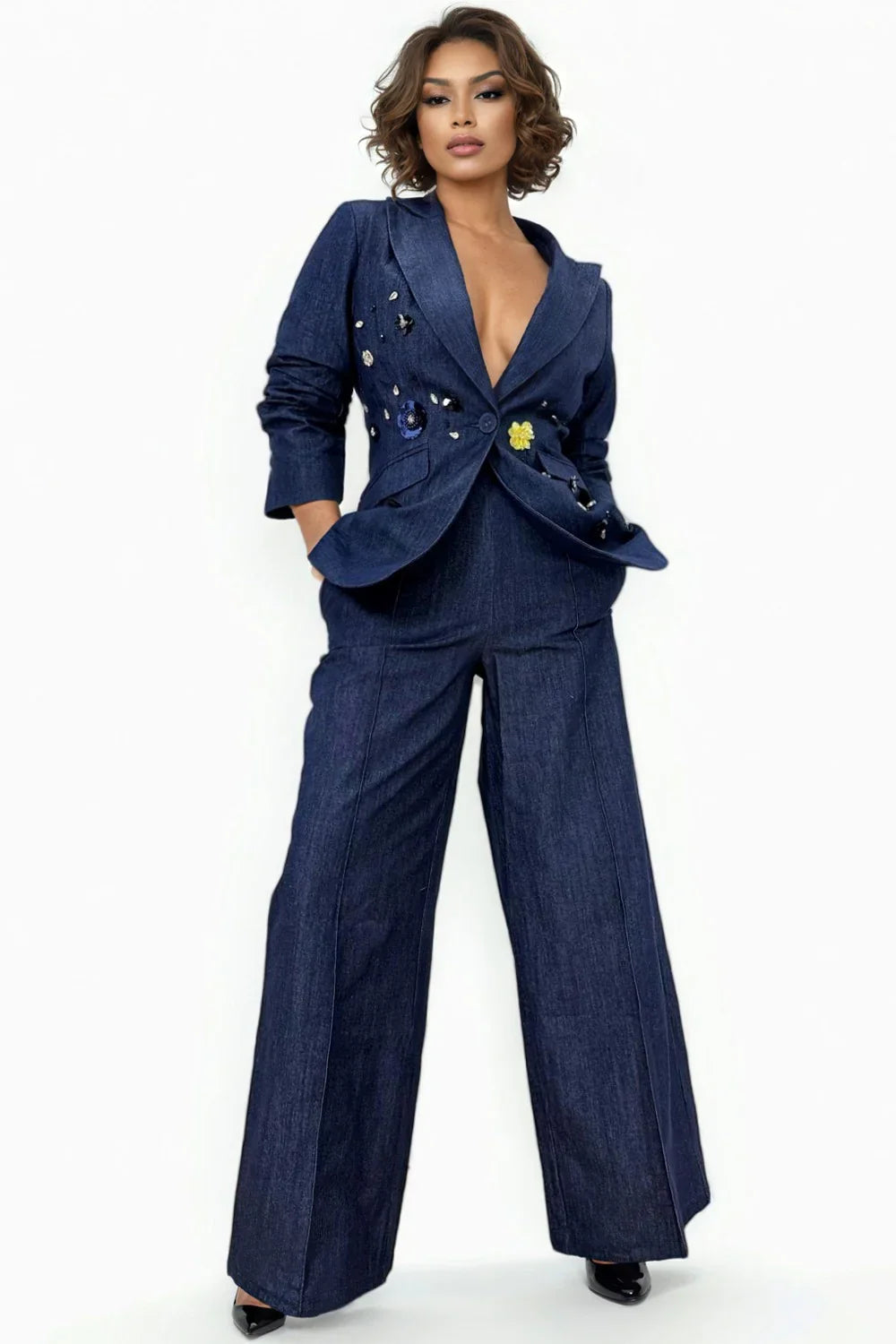 A woman modeling a denim blue ruched sleeve pant suit with a jacket and pants, standing against a white background, denim jacket has yellow & dark blue flowers.