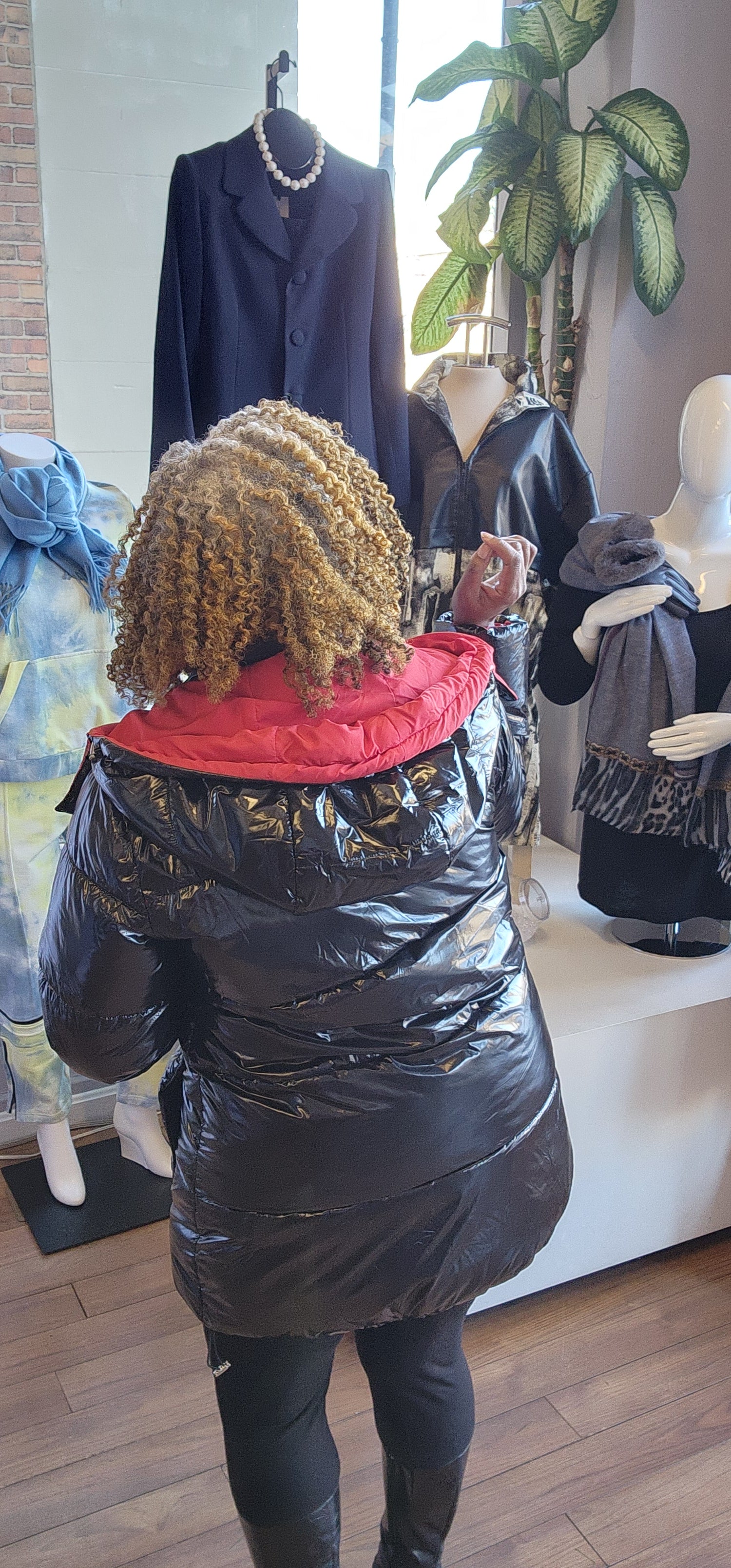 A person standing in a retail environment wearing a sleek black and red puffer coat with a removable hoodie. The coat has a dual-zipper design and the person is also wearing black boots.