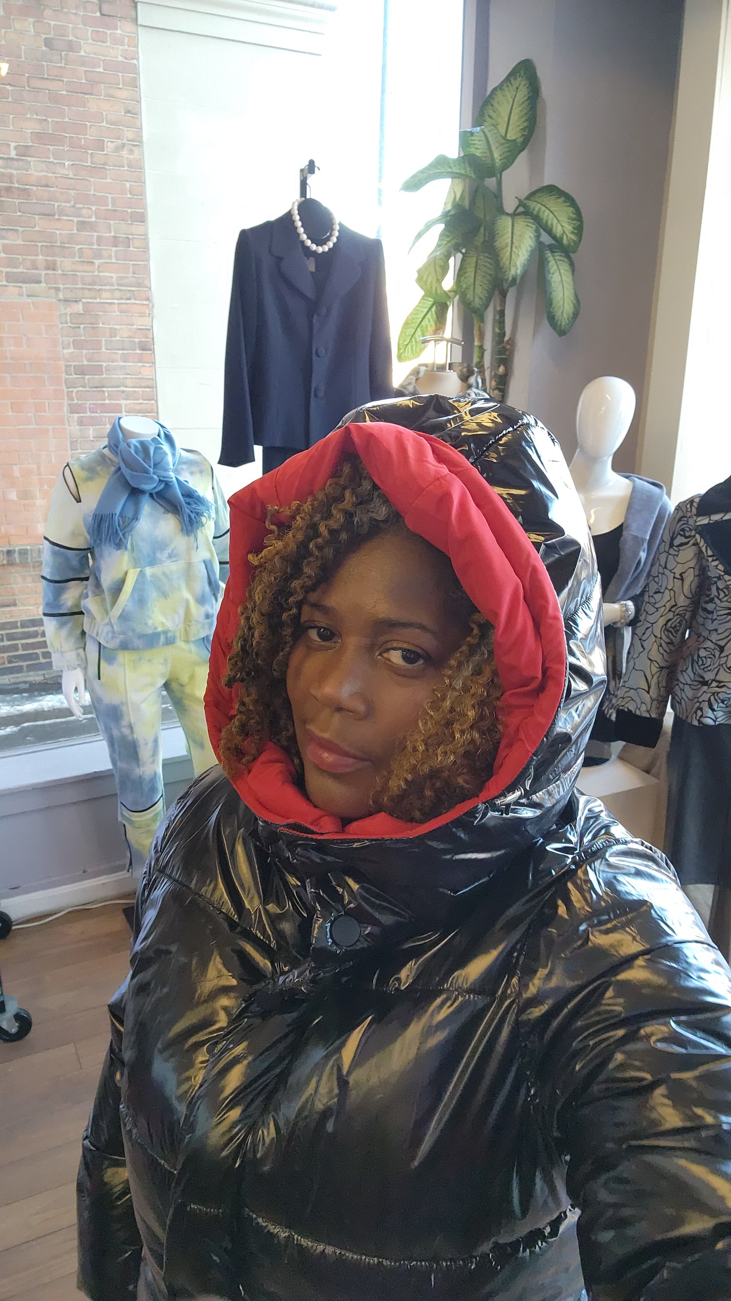 A person standing in a retail environment wearing a sleek black and red puffer coat with a removable hoodie. The coat has a dual-zipper design and the person is also wearing black boots.