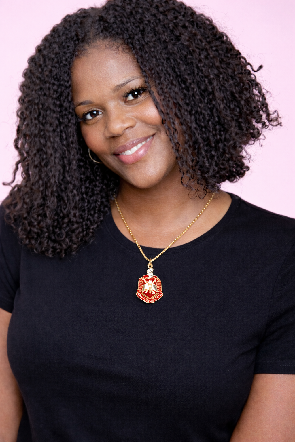 Woman wearing a black shirt and gold necklace with an DST pendant against a pink background