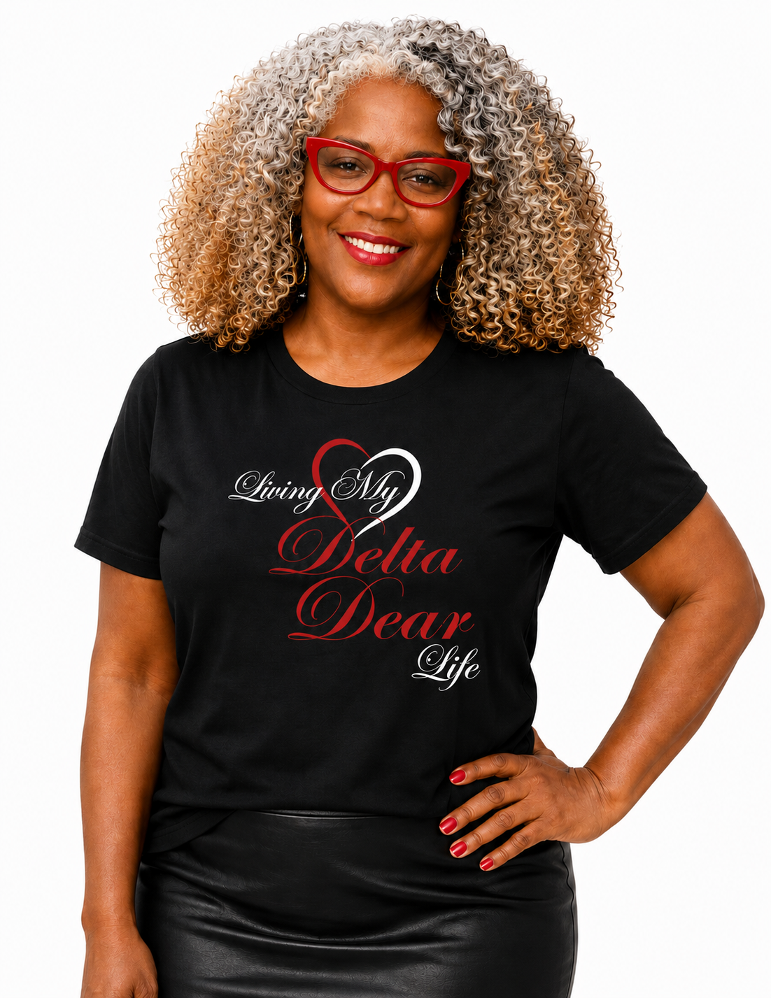 A smiling woman with curly gray-and-blonde hair and red glasses wears a black graphic T-shirt with the phrase 'Living My Delta Dear Life' in white and red script, paired with a black skirt, against a plain white background.