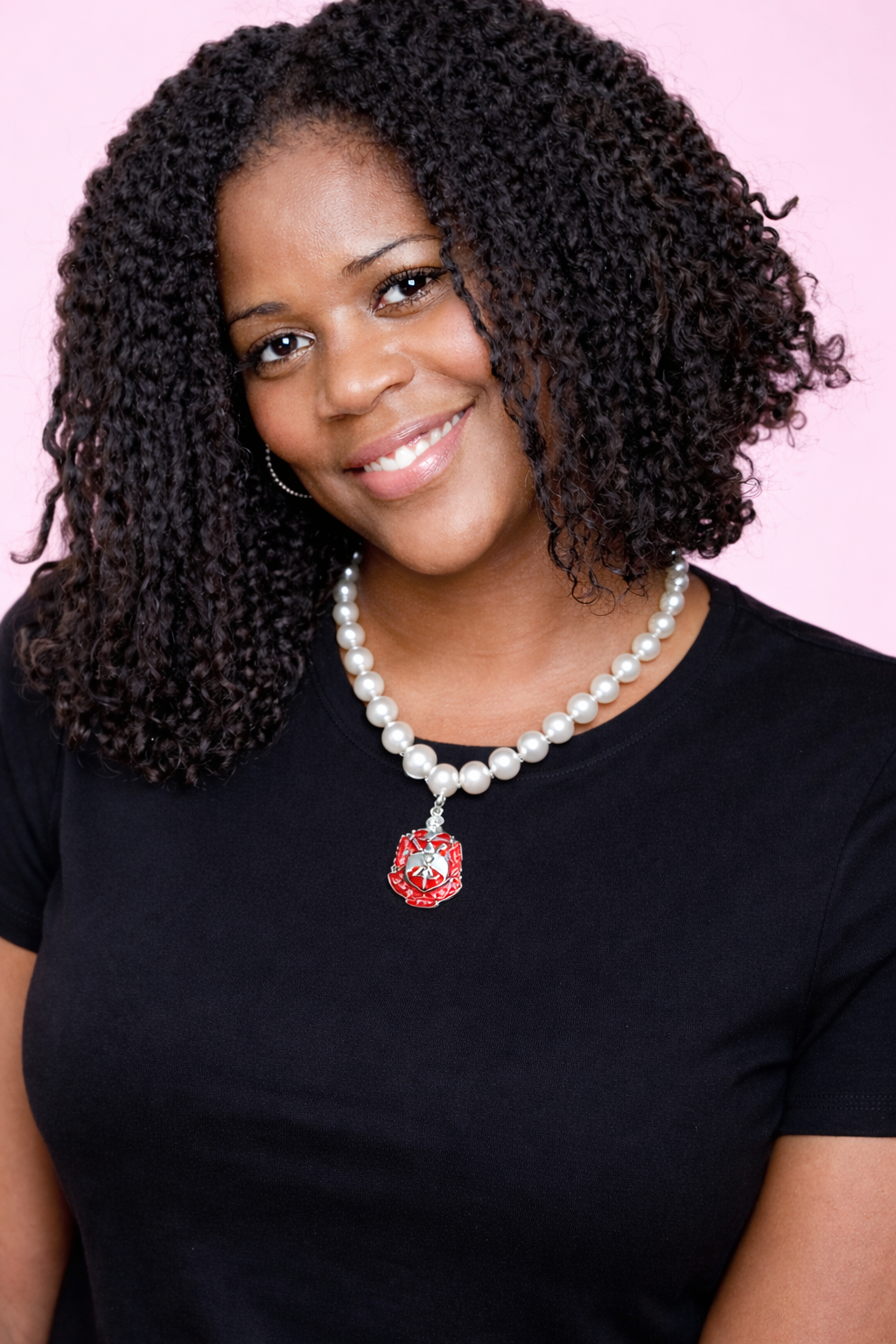 Woman wearing a black shirt with a red and gold emblem and pearl necklace with shield on a white background