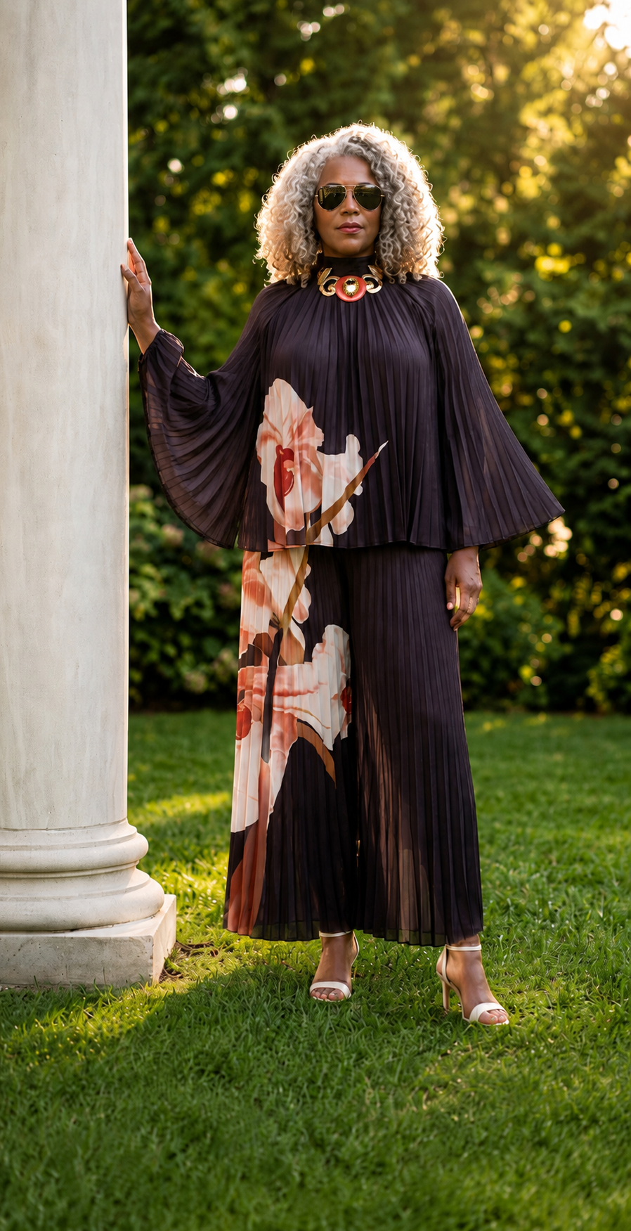A woman stands outdoors on a grassy lawn beside a white column, wearing a black pleated 2 piece wide-leg pant set with a large floral print in pink, peach, and cream. She is also wearing sunglasses and peach high-heel sandals.