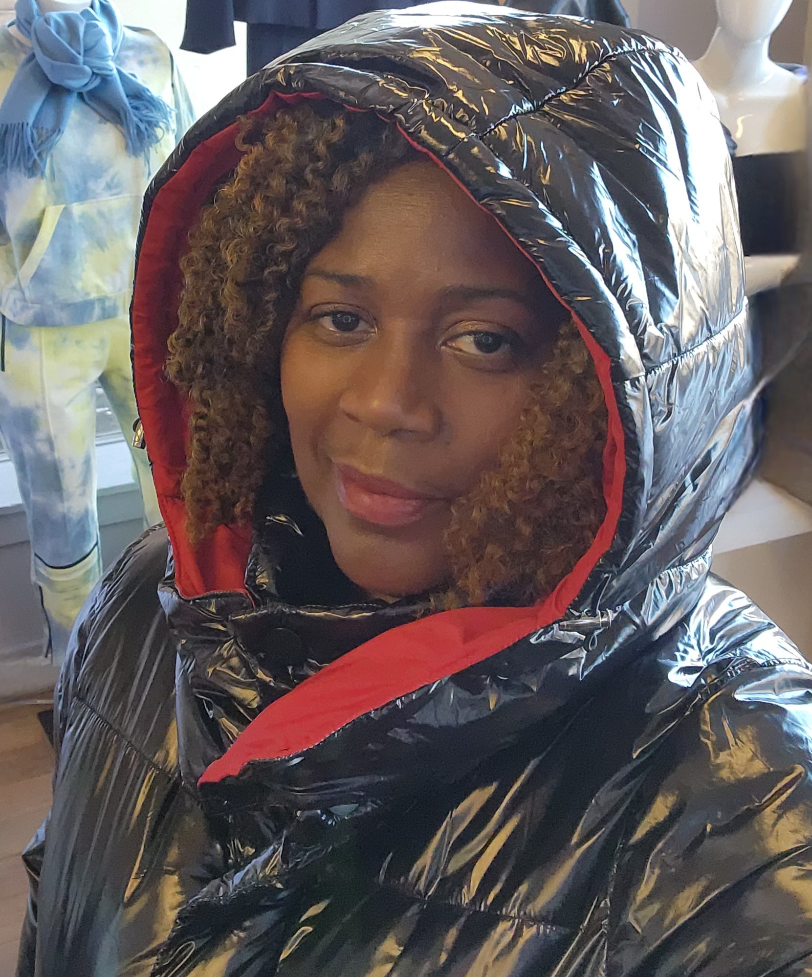 A person standing in a retail environment wearing a sleek black and red puffer coat with a removable hoodie. The coat has a dual-zipper design and the person is also wearing black boots.