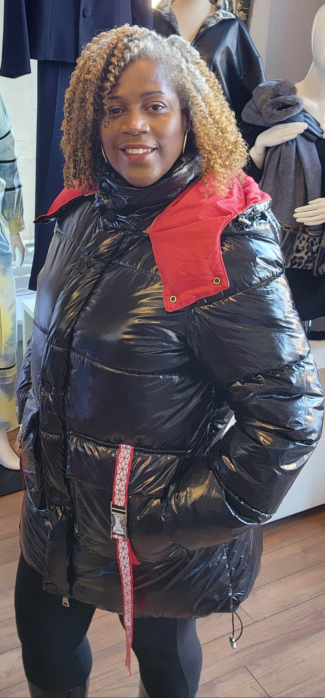 A person standing in a retail environment wearing a sleek black and red puffer coat with a removable hoodie. The coat has a dual-zipper design and the person is also wearing black boots.