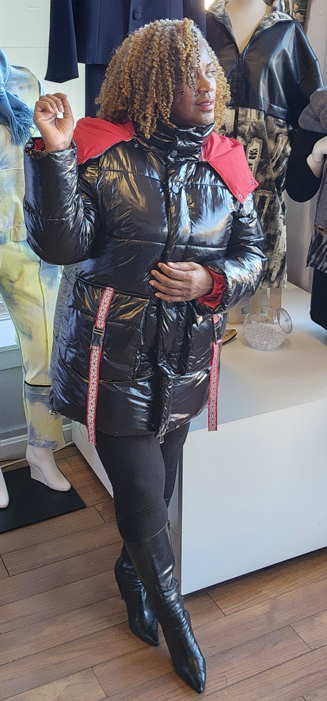A person standing in a retail environment wearing a sleek black and red puffer coat with a removable hoodie. The coat has a dual-zipper design and the person is also wearing black boots.