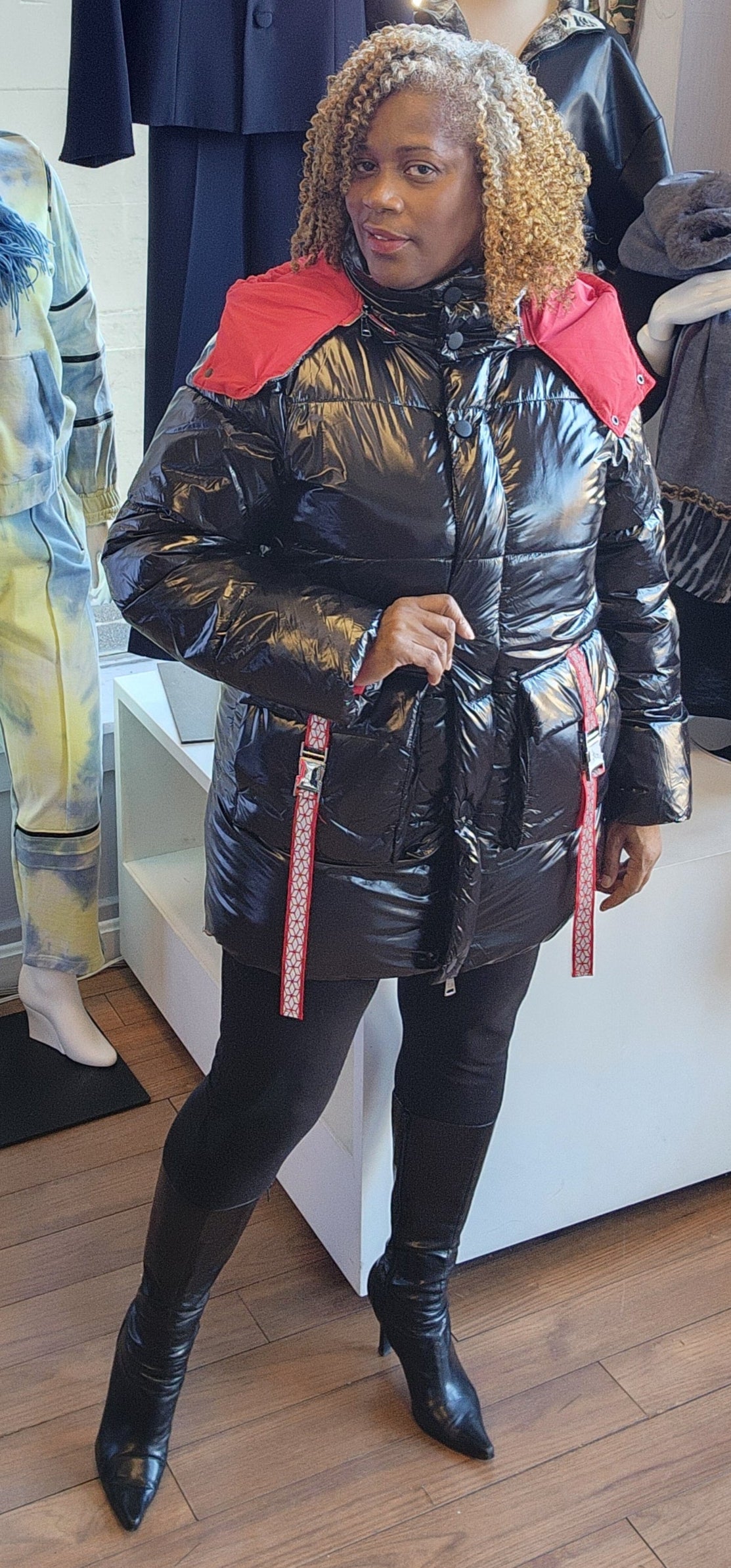 A person standing in a retail environment wearing a sleek black and red puffer coat with a removable hoodie. The coat has a dual-zipper design and the person is also wearing black boots.