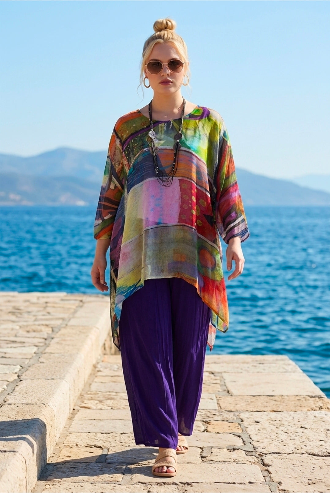 Woman wearing a colorful shirt and purple pants standing on a stone pathway by the sea.