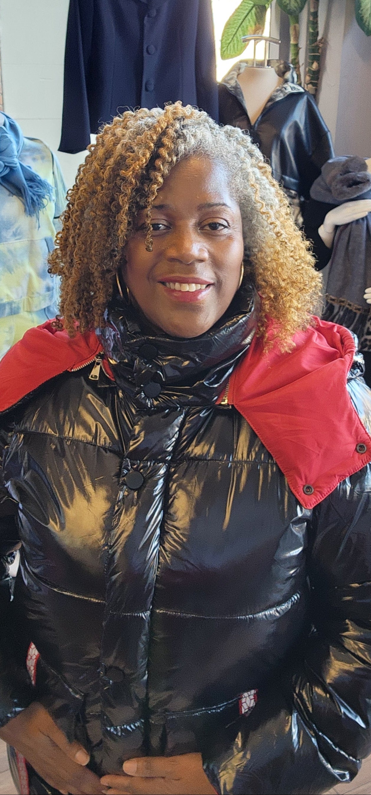A person standing in a retail environment wearing a sleek black and red puffer coat with a removable hoodie. The coat has a dual-zipper design and the person is also wearing black boots.